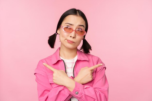 Keuze Stijlvol Koreaans meisje Aziatisch vrouwelijk model wijst vingers zijwaarts toont twee varianten productadvertentie die items demonstreert die over roze achtergrond staan