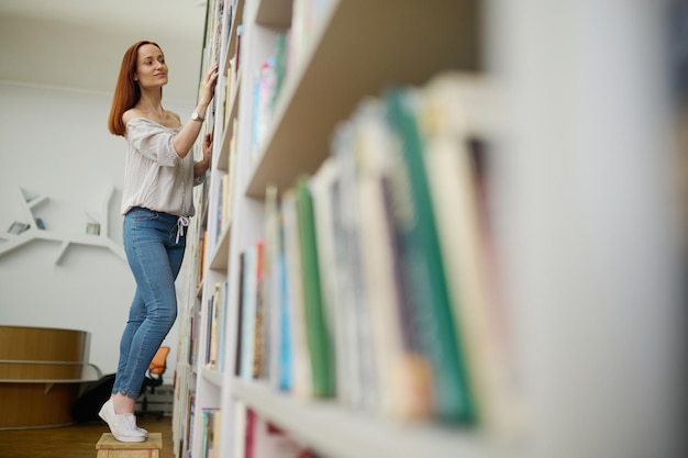 Kies boek. Jonge slanke langharige lachende vrouw in blouse en spijkerbroek op zoek naar boek staande handen aanraken boekenplank in bibliotheek