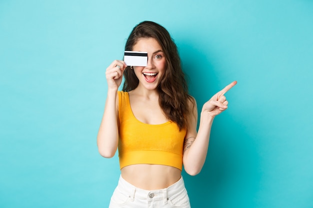 Kijk daar. Stijlvolle moderne vrouw die plastic creditcard toont, glimlacht en naar rechts wijst, wegwijst naar banner of logo, staande tegen een blauwe achtergrond