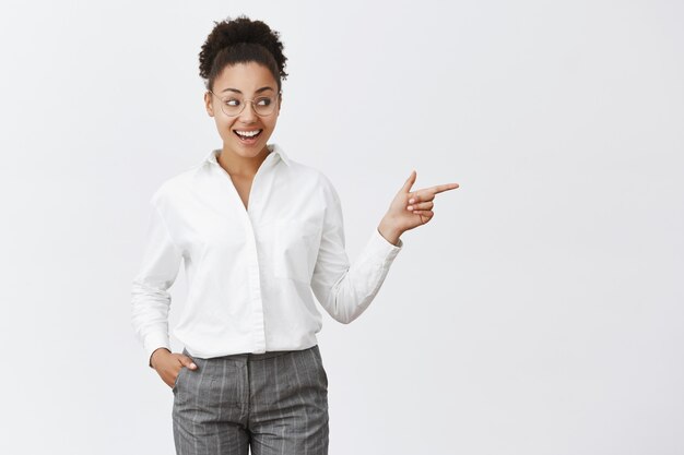 Kijk eens meid. Portret van verbaasd en blij goed uitziende gelukkige donkere vrouw in transparante bril, hand in zak vasthoudend en met een lichte glimlach naar rechts wijzend, onder de indruk