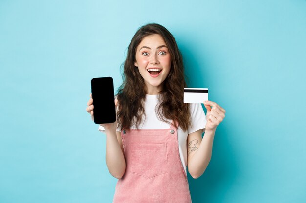 Kijk hier. Opgewonden jong schattig meisje met leeg mobiel scherm en plastic creditcard, kijk verbaasd naar de camera, staande over blauwe achtergrond.