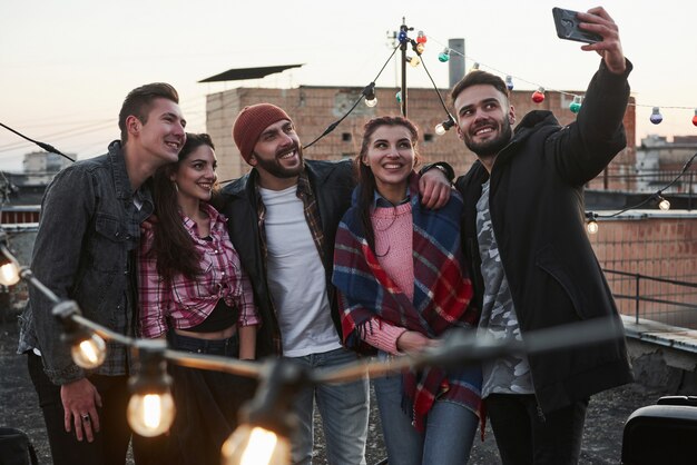 Kijk in de telefoon. Groep jonge vrolijke vrienden met plezier, omhelzen elkaar en neemt selfie op het dak met gloeilampen versieren