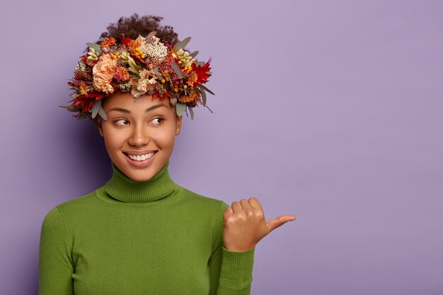 Kijk opzij. Vrolijke tedere krullende vrouw draagt herfstkrans rond het hoofd, wijst weg op kopie ruimte, lacht aangenaam