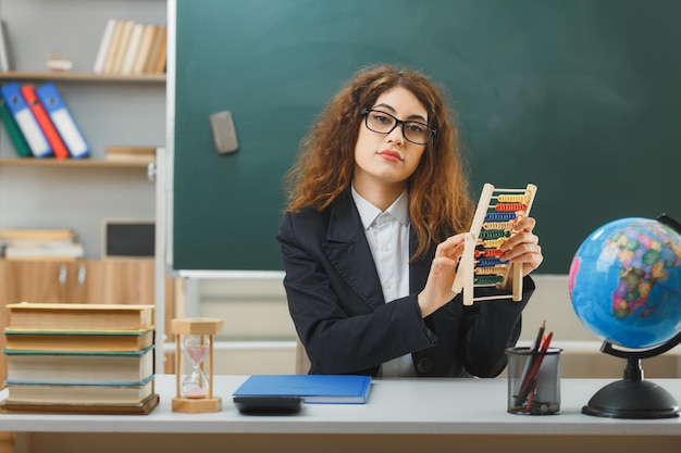 Kijken naar camera jonge vrouwelijke leraar die een bril draagt en een telraam vasthoudt die aan een bureau zit met schoolhulpmiddelen in de klas