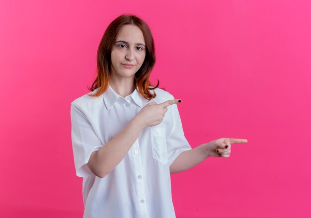 Kijkend naar camera jonge roodharige meisje wijst naar kant geïsoleerd op roze achtergrond met kopie ruimte