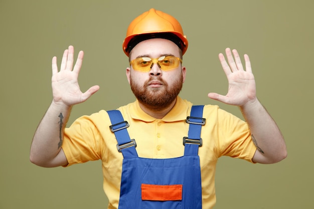 Gratis foto kijkend naar camera spreidende handen jonge bouwer man in uniform geïsoleerd op groene achtergrond