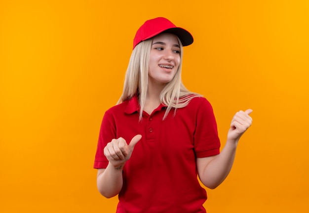 Kijkend naar de achterkant lachende levering jonge vrouw, gekleed in rode t-shirt en pet in tandsteun wijst op de rug op geïsoleerde oranje muur