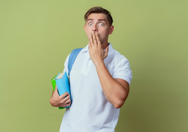 Kijkend naar de camera bang jonge knappe mannelijke student draagt rugtas met boeken en bedekte mond met hand geïsoleerd op olijfgroene achtergrond