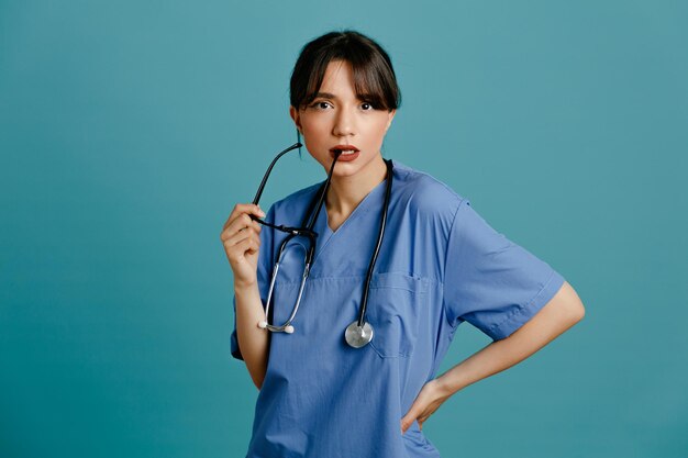 Gratis foto kijkend naar de camera die de hand op de heupen zet, jonge vrouwelijke arts die een uniforme stethoscoop draagt, geïsoleerd op een blauwe achtergrond