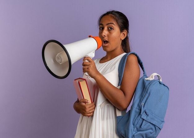 Kijkend naar de camera jong schoolmeisje draagt rugtas met boeken en spreekt op luidspreker op geïsoleerde paarse achtergrond