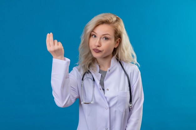 Kijkend naar de camera jonge arts die een stethoscoop in medische jurk draagt die contant gebaar op blauwe muur toont