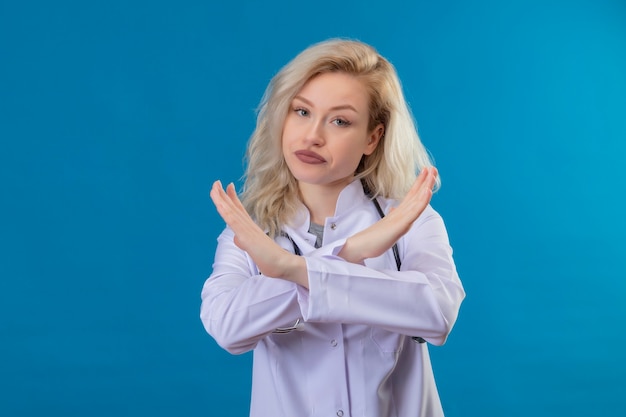 Kijkend naar de camera jonge arts die een stethoscoop in medische jurk draagt, doet geen gebaar op de blauwe muur