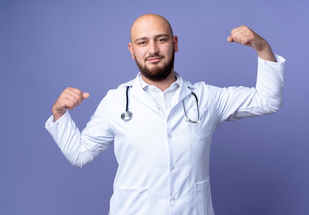 Kijkend naar de camera jonge kale mannelijke arts die medische mantel en een stethoscoop draagt, doet sterk gebaar geïsoleerd op blauwe achtergrond
