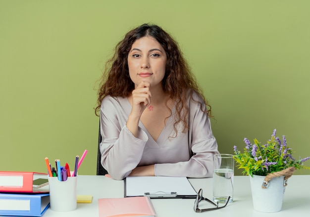 Kijkend naar de camera jonge mooie vrouwelijke kantoormedewerker zittend aan een bureau met kantoorhulpmiddelen zetten hand onder de kin geïsoleerd op olijf achtergrond