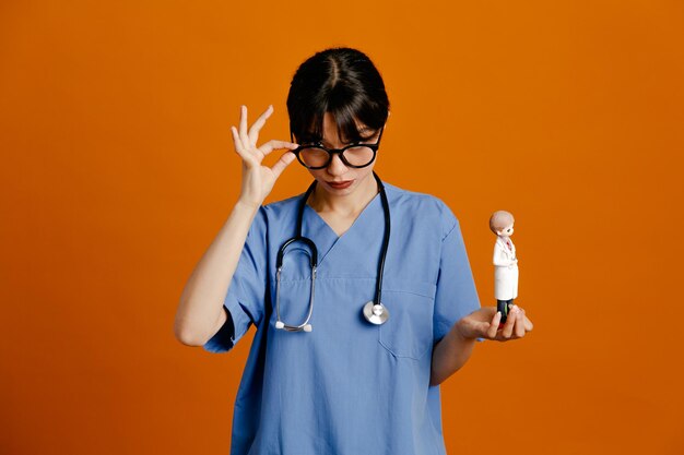 Kijkend naar de camera met speelgoed jonge vrouwelijke arts met een uniforme stethoscoop geïsoleerd op een oranje achtergrond