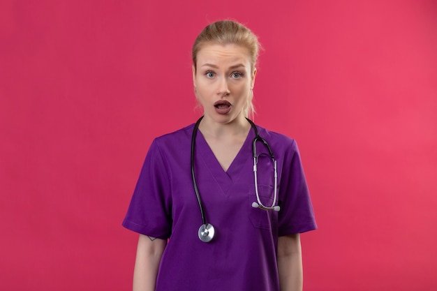 Kijkend naar de camera verbaasde jonge arts die paarse medische jurk in een stethoscoop op geïsoleerde roze muur draagt
