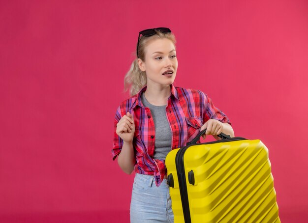 Kijkend naar de jonge vrouwelijke reiziger die aan de zijkant een rood shirt en een bril op haar hoofd draagt en een koffer op geïsoleerde roze muur houdt