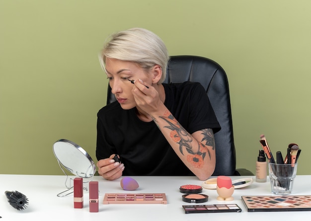 Kijkend naar de spiegel zit een mooi meisje aan tafel met make-uptools, teken een pijl met eyeliner geïsoleerd op een olijfgroene muur