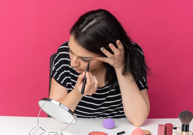 Kijkend naar de spiegel zit een mooi meisje aan tafel met make-uptools, teken een pijl met eyeliner geïsoleerd op een roze muur