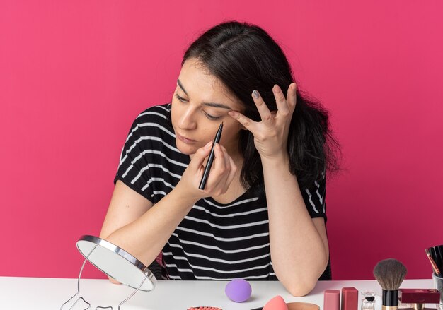 Kijkend naar de spiegel zit een mooi meisje aan tafel met make-uptools, teken een pijl met eyeliner geïsoleerd op een roze muur