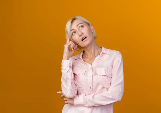 Kijkend naar denkende jonge blonde Slavische vrouw die vinger op hoofd zet dat op oranje muur wordt geïsoleerd