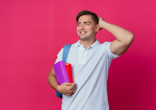 Kijkend naar een glimlachende jonge, knappe mannelijke student die een rugtas draagt die boeken vasthoudt en de hand op het hoofd legt