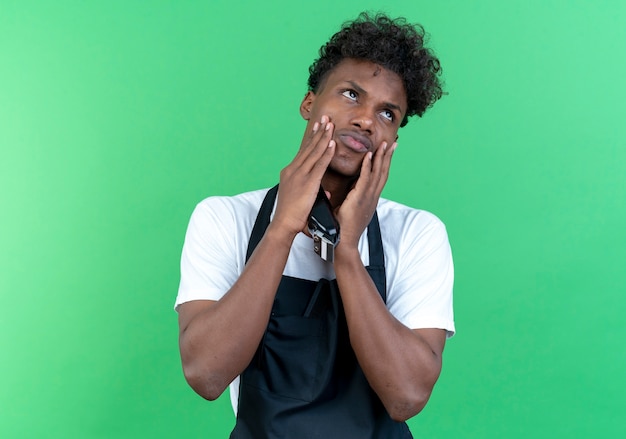 Kijkend naar een trieste jonge Afro-Amerikaanse mannelijke kapper die een uniform draagt met een tondeuse en handen op de wangen legt