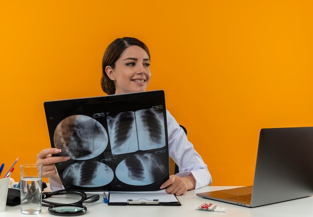 Kijkend naar kant amiling jonge vrouwelijke arts die medische mantel draagt met een stethoscoop zittend aan een bureau werkt op de computer met medische hulpmiddelen die röntgenfoto op isolatie gele muur houdt