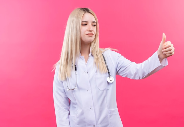 Gratis foto kijkend naar kant arts jonge blonde meisje draagt ?? een stethoscoop in medische jurk haar duim omhoog op geïsoleerde roze muur