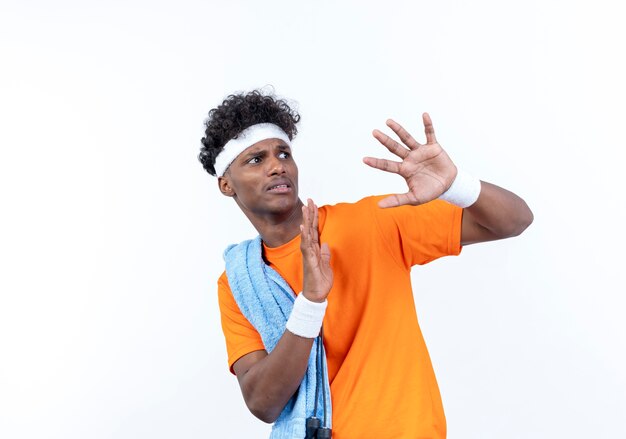 Kijkend naar kant bang jonge Afro-Amerikaanse sportieve man met hoofdband en polsband spreidt handen met handdoek op schouder geïsoleerd op witte achtergrond