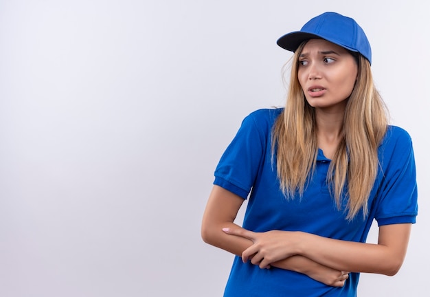 Kijkend naar kant betrokken jonge levering meisje dragen blauwe uniform en pet kruising handen geïsoleerd op een witte muur met kopie ruimte