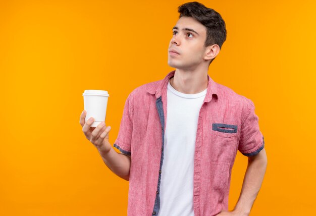 Kijkend naar kant blanke jonge man met roze shirt met kopje koffie op geïsoleerde oranje muur