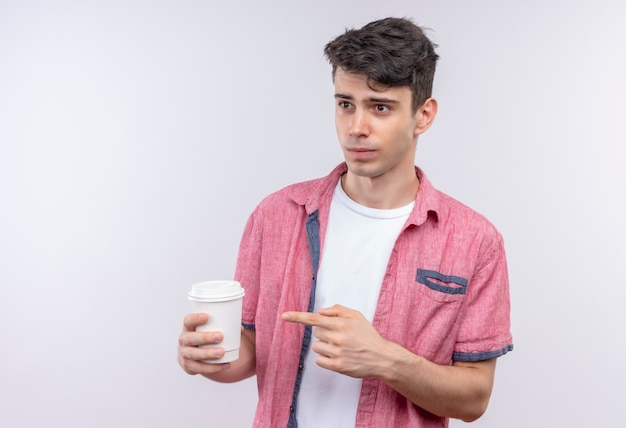 Kijkend naar kant blanke jonge man met roze shirt wijst naar kopje koffie op geïsoleerde witte muur