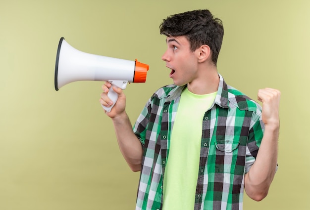 Kijkend naar kant blanke jongeman met groen shirt spreekt door luidspreker met ja gebaar op geïsoleerde groene muur