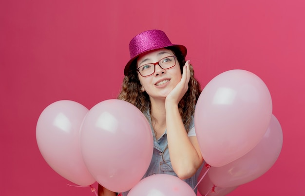 Kijkend naar kant blij jong meisje met bril en roze hoed staande achter ballonnen en hand op wang geïsoleerd op roze achtergrond