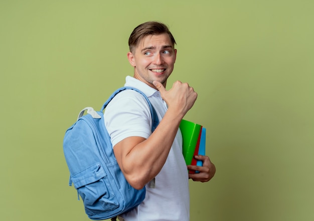 Kijkend naar kant glimlachende jonge knappe mannelijke student die rugtas draagt die boeken en punten aan kant draagt die op olijfgroene muur wordt geïsoleerd