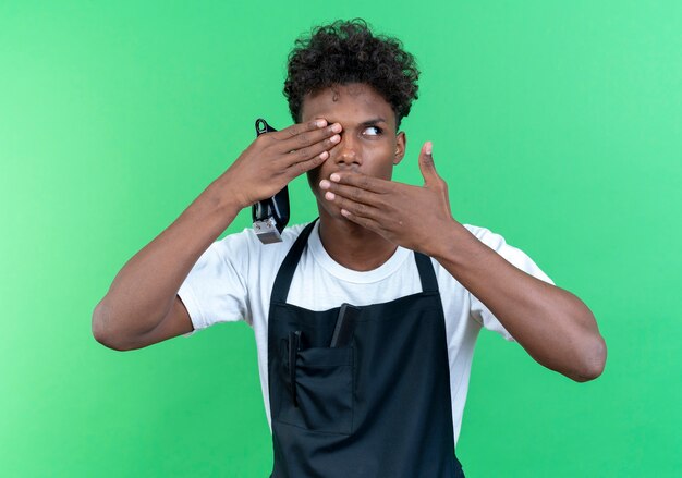 Kijkend naar kant jonge Afro-Amerikaanse mannelijke kapper dragen van uniforme haar tondeuse bedekte mond en oog met handen geïsoleerd op groene muur