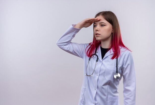 Kijkend naar kant jonge dokter meisje dragen stethoscoop medische mantel kijken afstand met hand op geïsoleerde witte achtergrond met kopie ruimte