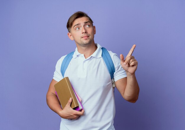 Kijkend naar kant jonge knappe mannelijke student die rugtas draagt die boeken en punten aan kant houdt die op blauwe muur met exemplaarruimte wordt geïsoleerd