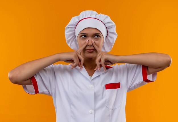 Kijkend naar kant jonge kok vrouwelijke chef-kok uniforme gesloten neus op geïsoleerde oranje muur dragen