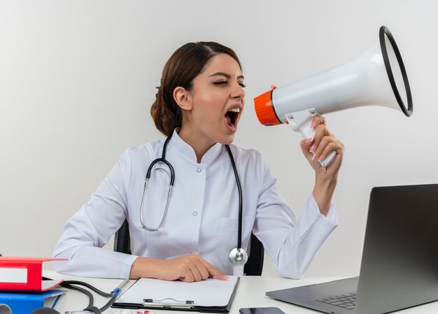 Kijkend naar kant jonge vrouwelijke arts die medische mantel draagt met een stethoscoop zittend aan een bureau werkt op de computer met medische hulpmiddelen spreekt op luidspreker op geïsoleerde witte muur met kopie ruimte