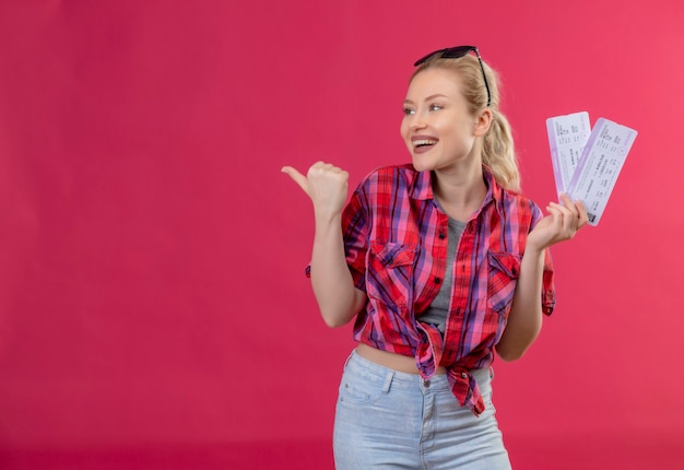 Kijkend naar kant jonge vrouwelijke reiziger die rood shirt in glazen draagt die kaartjes op geïsoleerde roze muur houdt