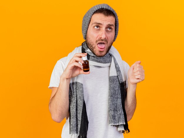 Kijkend naar kant jonge zieke man met winter muts en sjaal met geneeskunde in glazen fles geïsoleerd op gele achtergrond