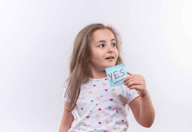 Kijkend naar kant klein schoolmeisje die wit t-shirt dragen die document teken op geïsoleerde witte muur houden