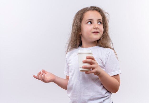 Kijkend naar kant klein schoolmeisje die witte t-shirt dragen die kop van koffie op geïsoleerde witte muur houden