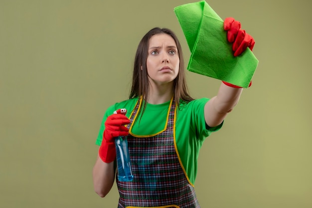 Kijkend naar kant schoonmaak jonge vrouw dragen uniform in rode handschoenen met schoonmaak spray met doek op geïsoleerde groene muur
