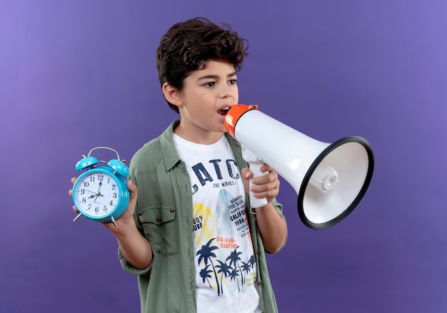 Kijkend naar kant spreekt kleine schooljongen op luidspreker en houdt wekker geïsoleerd op paarse muur