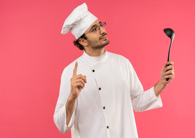Kijkend naar kant tevreden jonge mannelijke kok dragen uniforme chef-kok en glazen pollepel houden en wijst naar boven