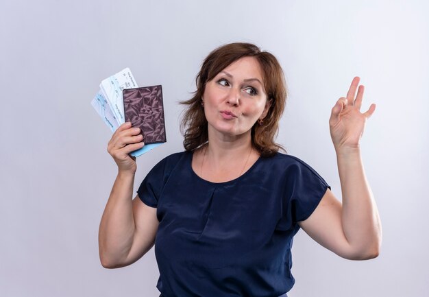 Kijkend naar kant tevreden reiziger vrouw met kaartjes en portemonnee van middelbare leeftijd met ok gebaar op geïsoleerde witte muur