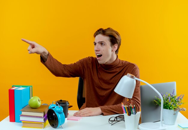 Kijkend naar kant verrast jonge student jongen zittend aan een bureau met schoolhulpmiddelen wijst naar boven